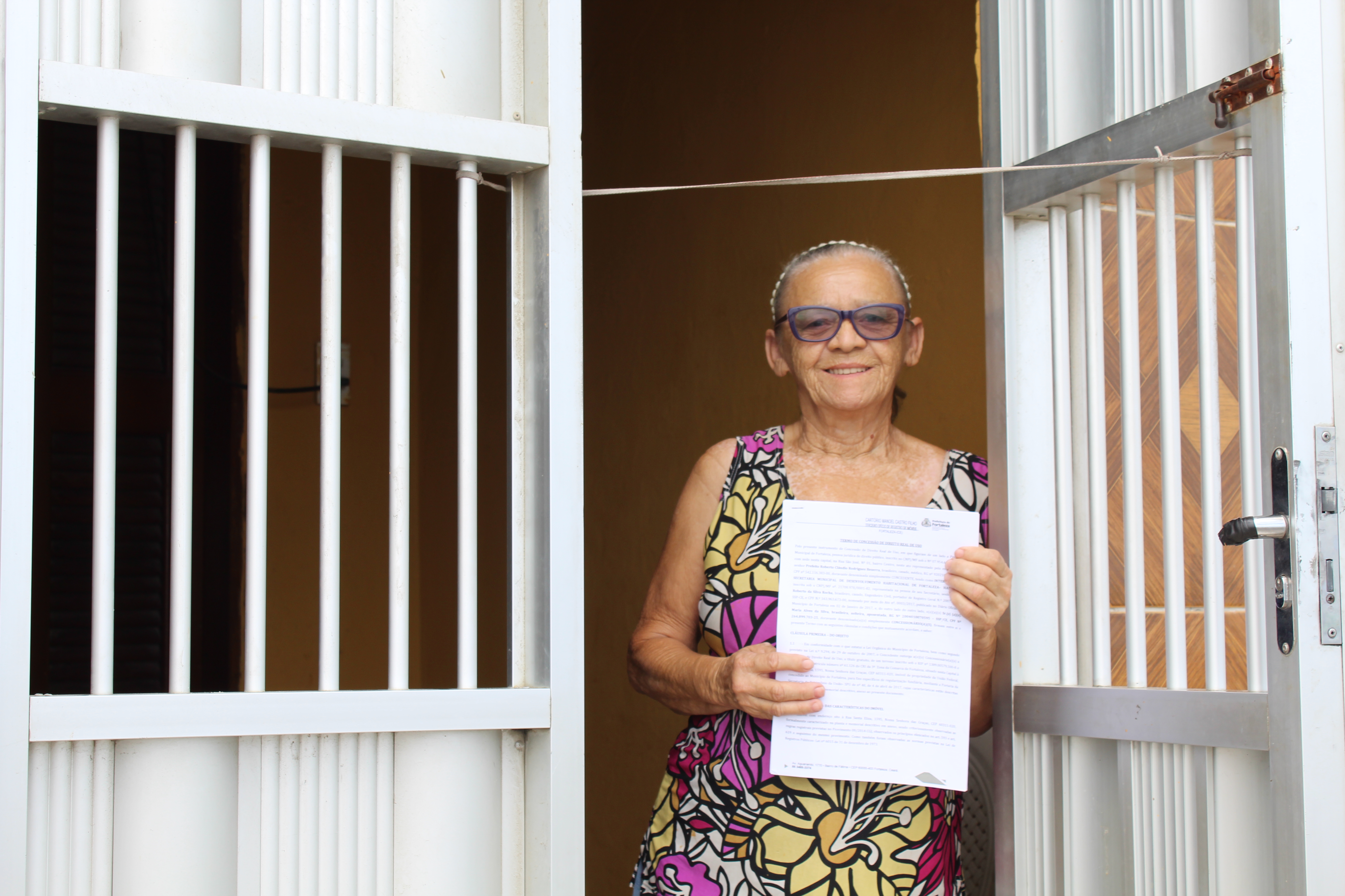 senhora de óculos posando para a foto na porta de casa e mostrando documento em mãos